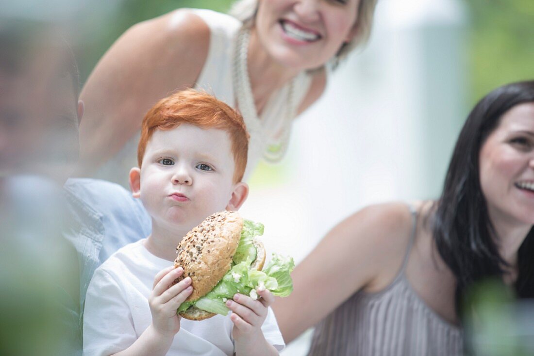 A young red-haired boy holding a sandwich surrounded by his family