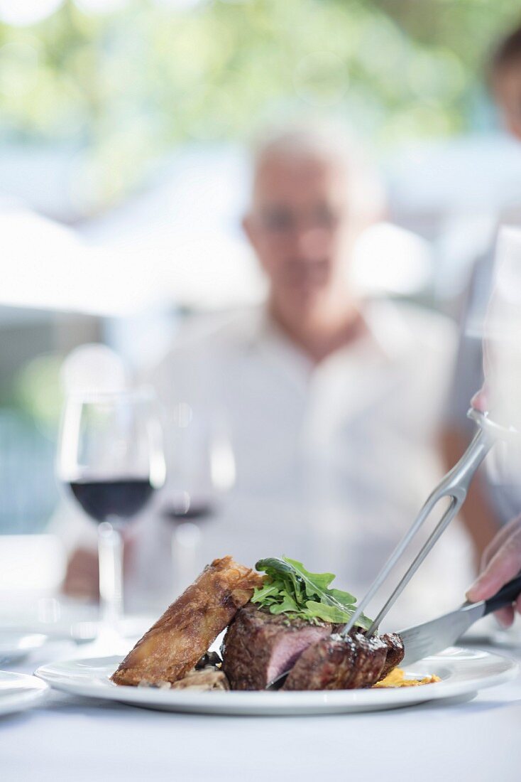 A meat dish and a glass of red wine on a summer table outside with a person in the background
