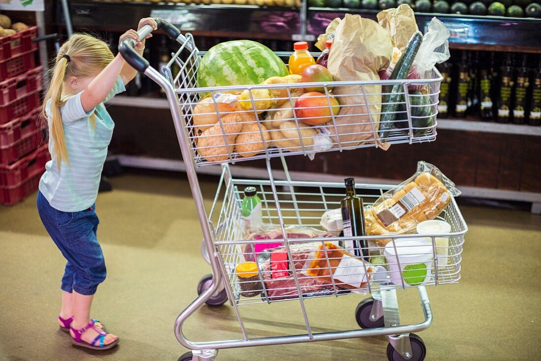 Kleines Mädchen schiebt vollen Einkaufswagen durch Supermarkt