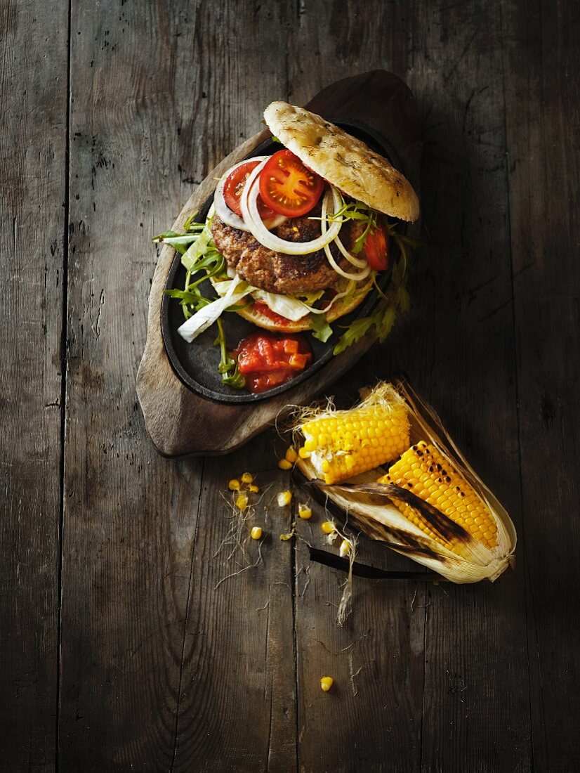 A hamburger with tomato salsa and grilled corn cobs