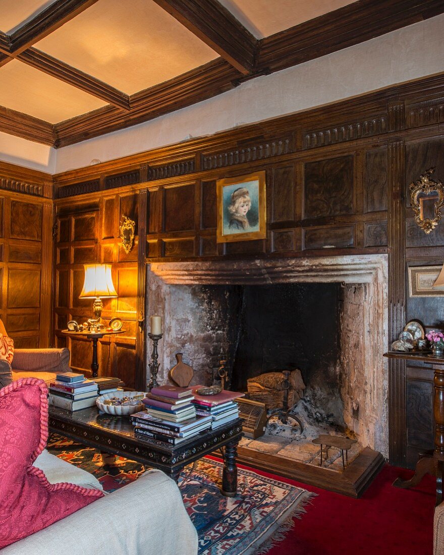 Wood panelling, coffered ceiling and books stacked on coffee table in front of large fireplace in classic interior