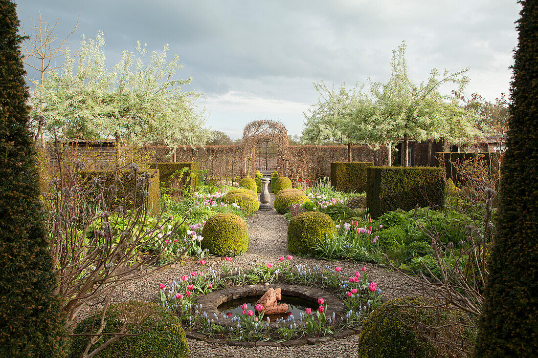 Brunnen mit Blumenrondell und Kiesweg mit geschnittenen Buchshecken in Gartenanlage