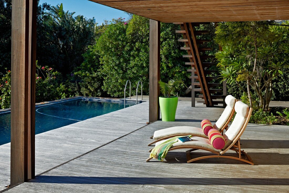 Sonnenliegen mit hellen Polstern und Rolle, auf grosszügiger Holzterrasse mit Überdachung, gegenüber Pool in tropischem Garten