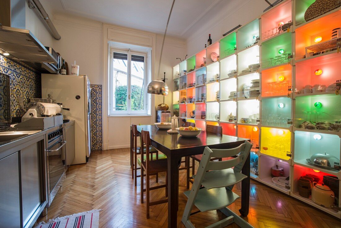 Dining area between stainless steel kitchen counter and colourful, illuminated shelving system holding crockery and kitchen utensils