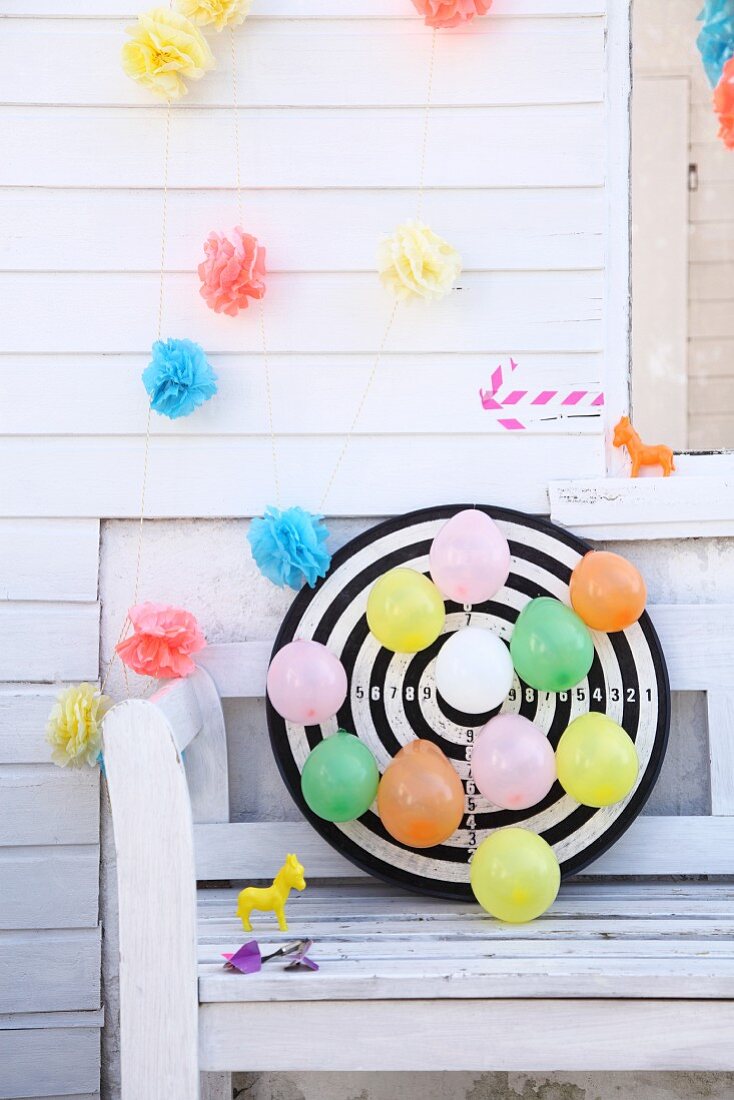 Colourful balloons pinned to dartboard below garlands of paper flowers on white wooden wall