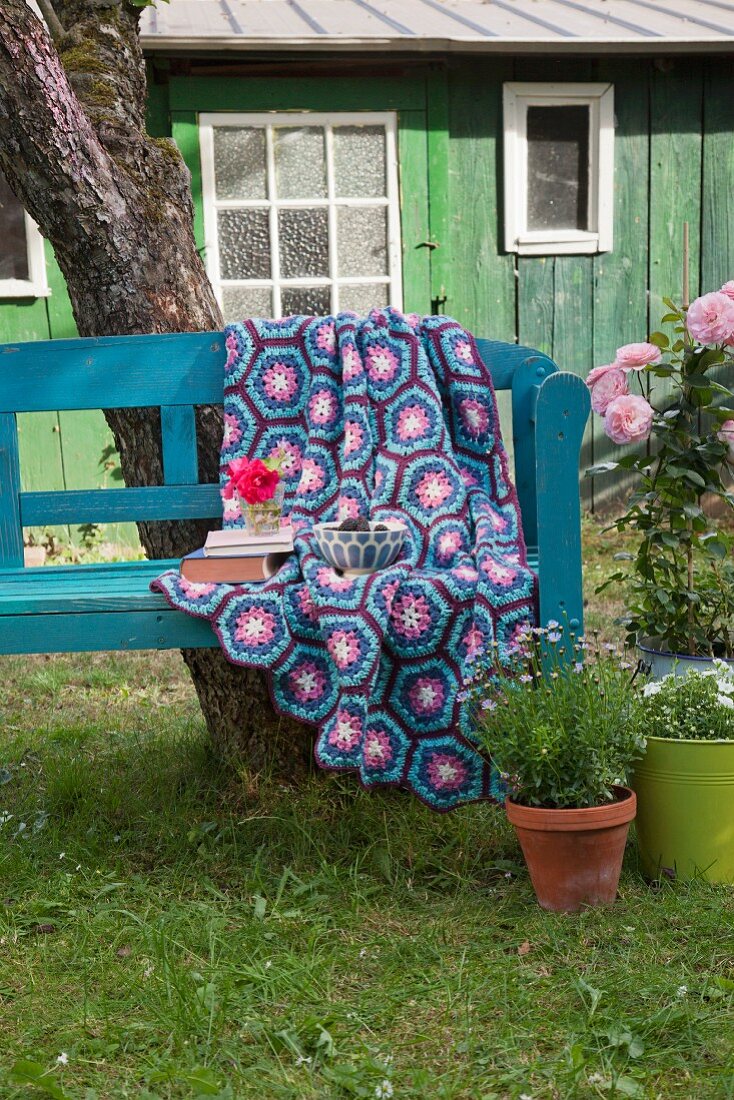 Häkeldecke aus Hexagons in Blau- und Violetttönen auf Gartenbank vor Hütte