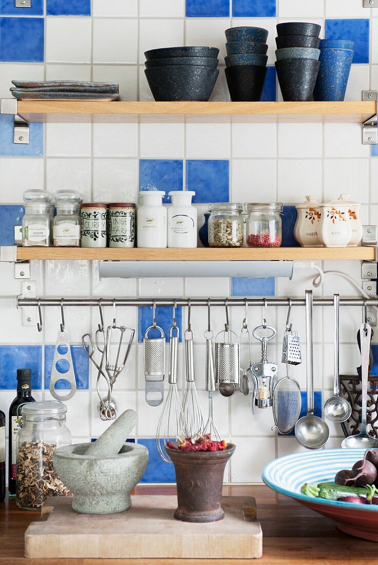 Shelves of spices and crockery above cooking utensils hung from stainless steel rod on kitchen wall with white and blue tiles