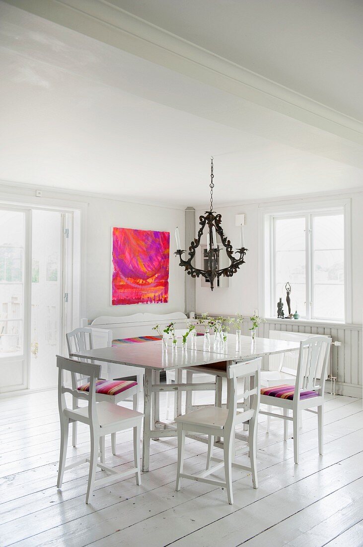 Weisser Tisch mit Holzstühlen unter Kronleuchter in ländlichem Ambiente mit weißem Dielenboden und modernem Bild