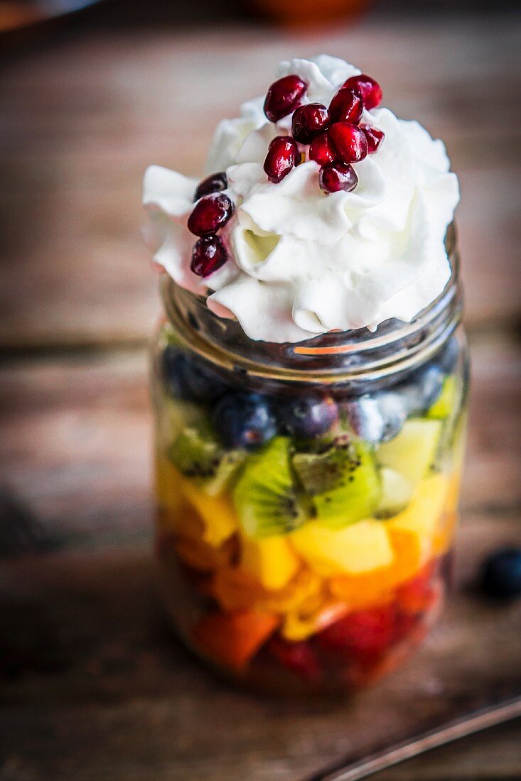 A colourful fruit salad in a jar topped with cream and pomegranate seeds