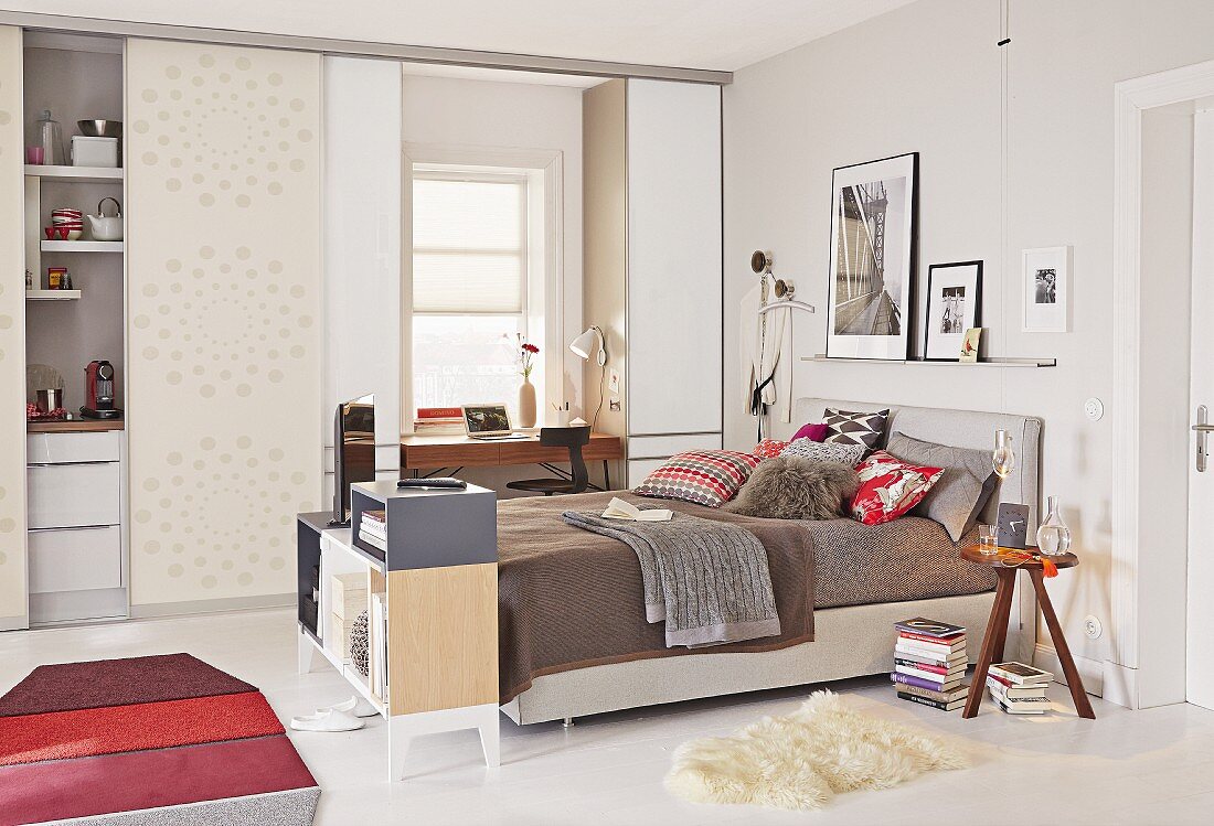 A box spring bed with a home office and a niche kitchen in the background separated by sliding doors in a one-room apartment