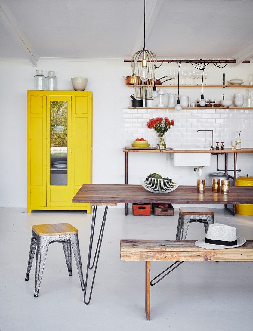 Schlichter Essplatz mit Klassiker Hocker vor Holztisch, im Hintergrund gelber Geschirrschrank neben Küchenzeile