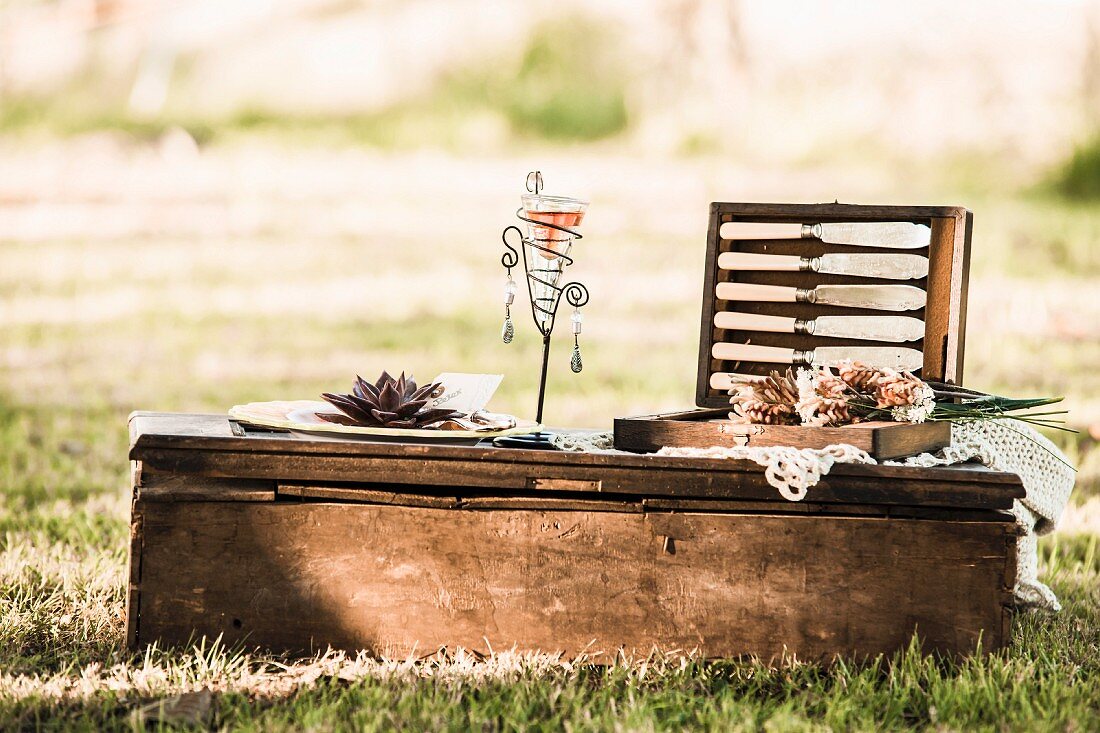 Dekoratives Windlicht und offener Picknick-Besteckkasten auf Vintage Bodentisch im Freien