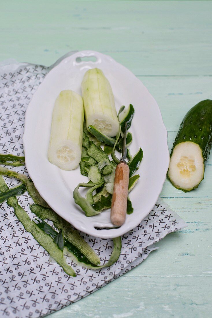 A peeled cucumber on a white porcelain plate with a peeler and peel