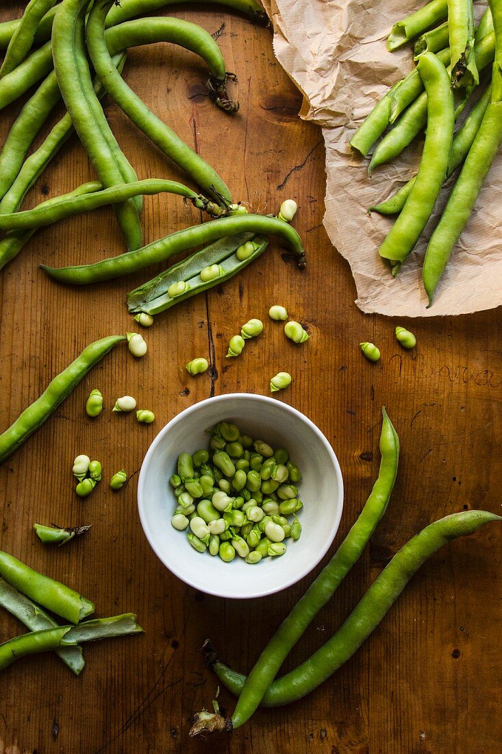A bowl of shelled broad beans surrounded by beans in pods