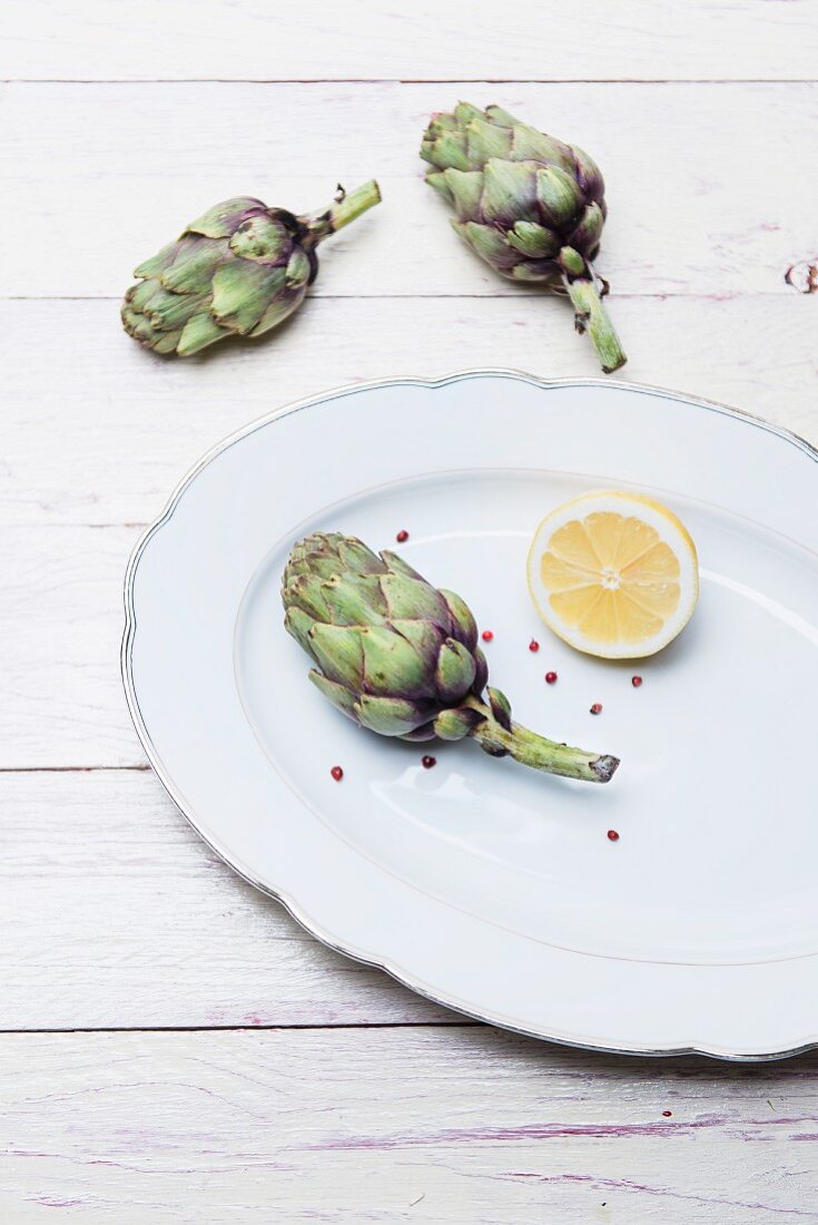 Fresh baby artichokes, half a lemon and pink peppercorns on a porcelain plate