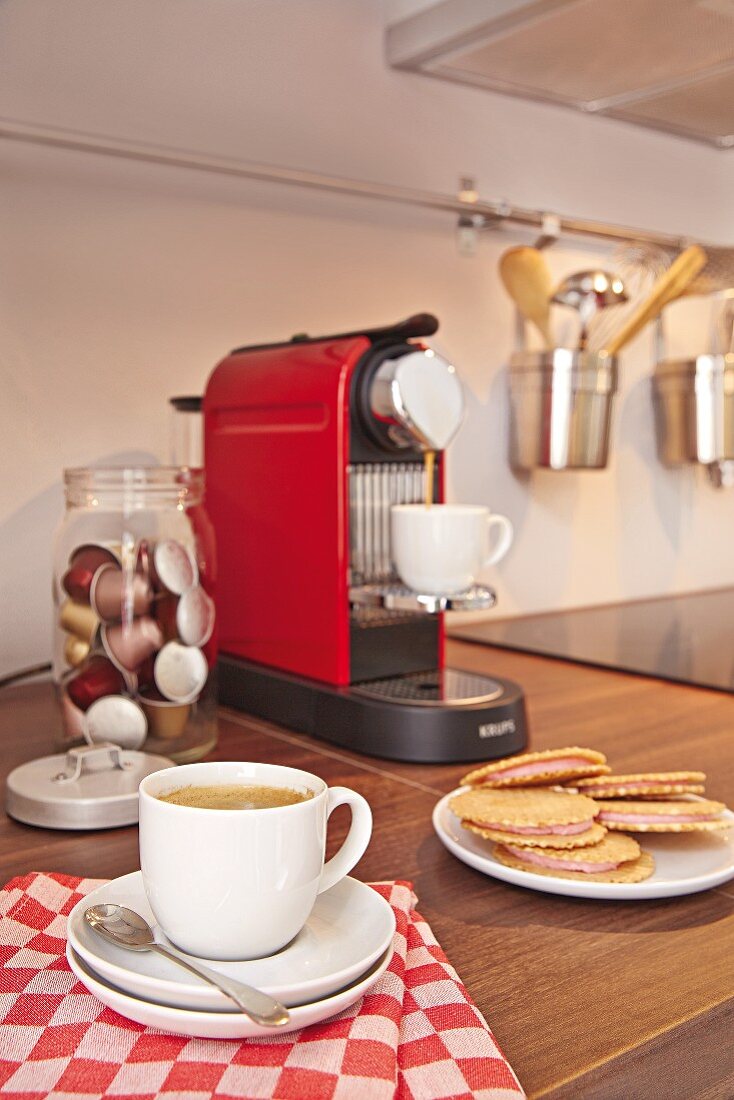 A mug of coffee on a checked tea towel and filled waffle biscuits with a coffee machine and a jar of coffee capsules in the background