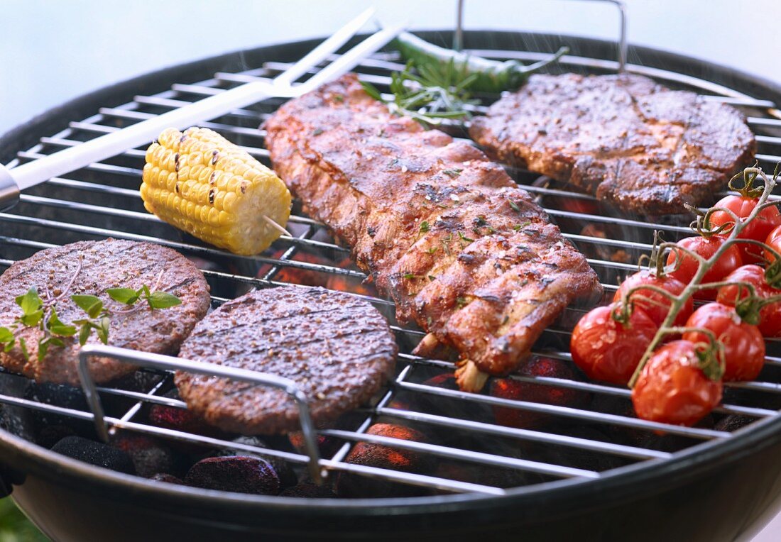 A beef steak, hamburgers, spare ribs, tomatoes and a corn cob on a barbecue
