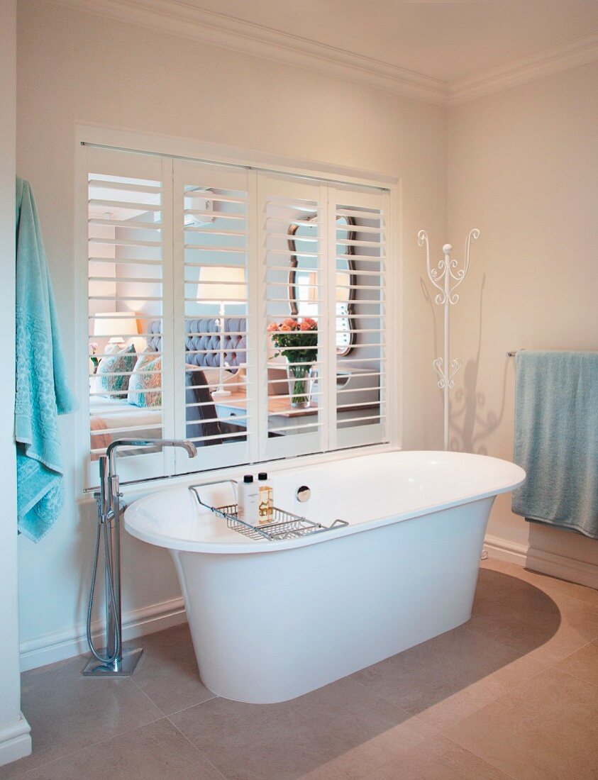 Free-standing bathroom with floor-mounted taps in front of interior window with open louvre shutters and view into bedroom