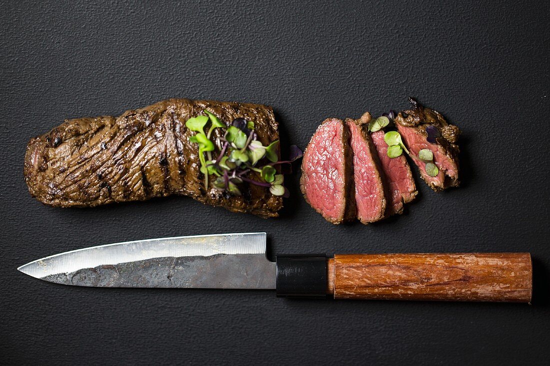 Sliced steak with shiso cress and a Japanese knife on a black surface