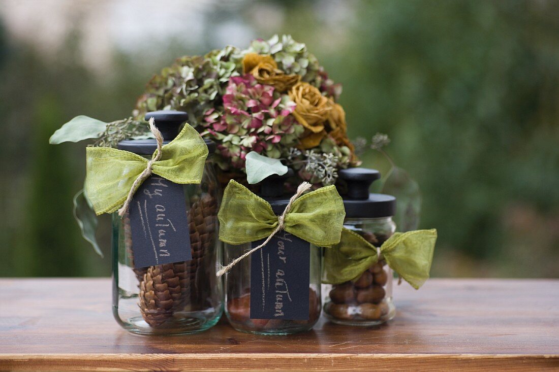 Nuts and pine cones in three storage jars with black lids and labels in front of posy of dried flowers