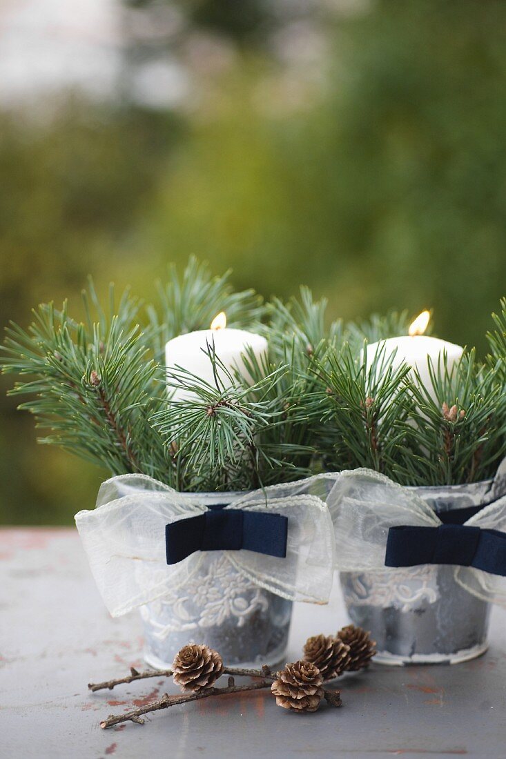 Two zinc buckets decorated with ribbon, spruce twigs and lit candles next to larch cones on surface