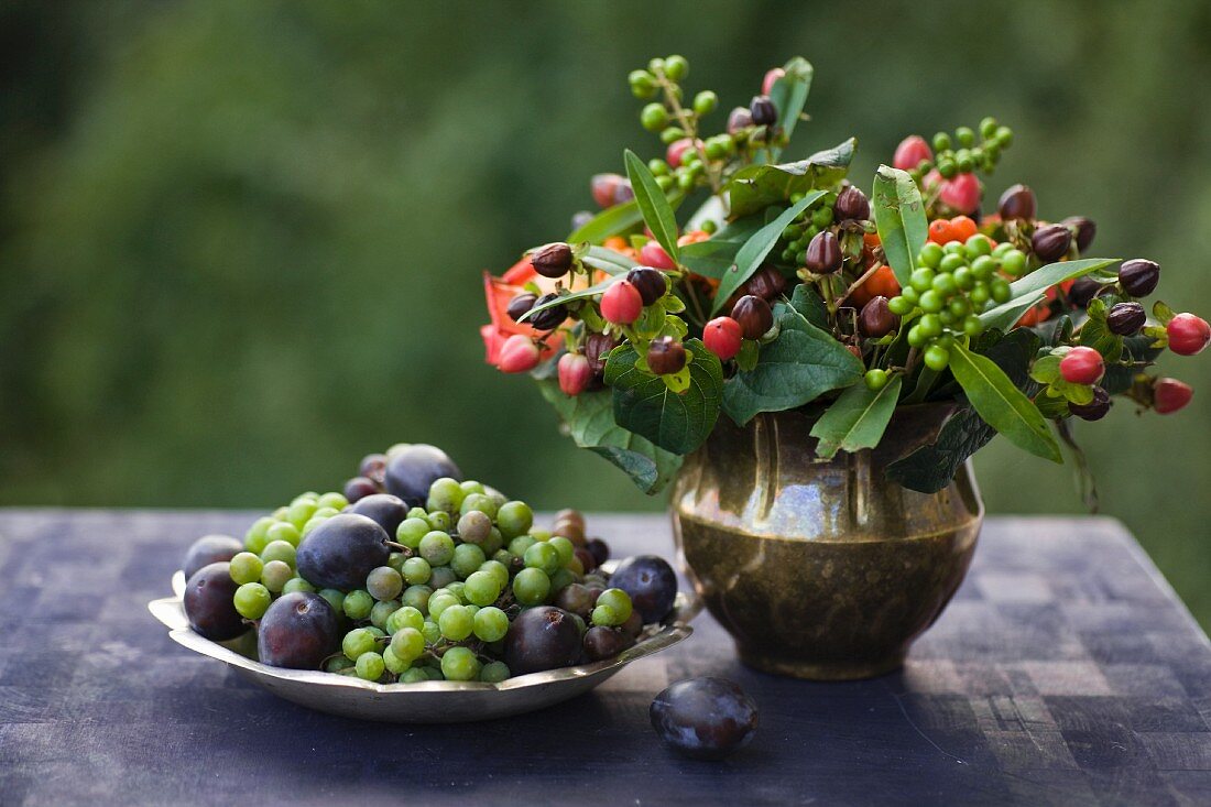 Herbststrauß mit Johanniskrautbeeren in einer Meesingvase, daneben Trauben und Pflaumen in einem Zinkteller