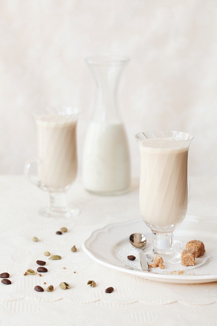 Zwei Gläser Gewürz-Milchkaffee mit Würfelzucker, Kaffeebohnen und Kardamomkapseln