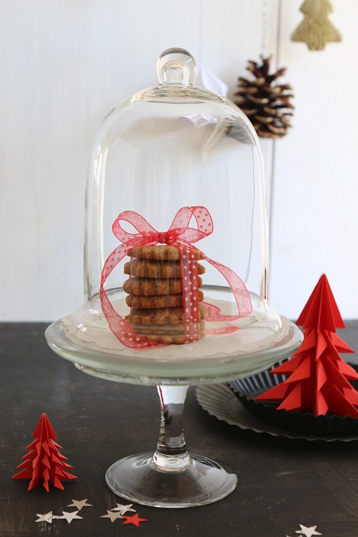 Stack of Christmas biscuits tied with ribbon under glass cover next to red paper Christmas tree
