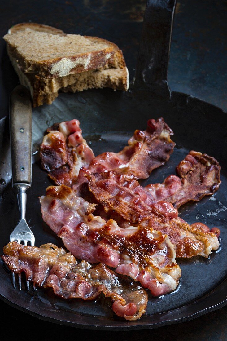 Gebratene Baconscheiben in Eisenpfanne mit Gabel und Brotscheiben