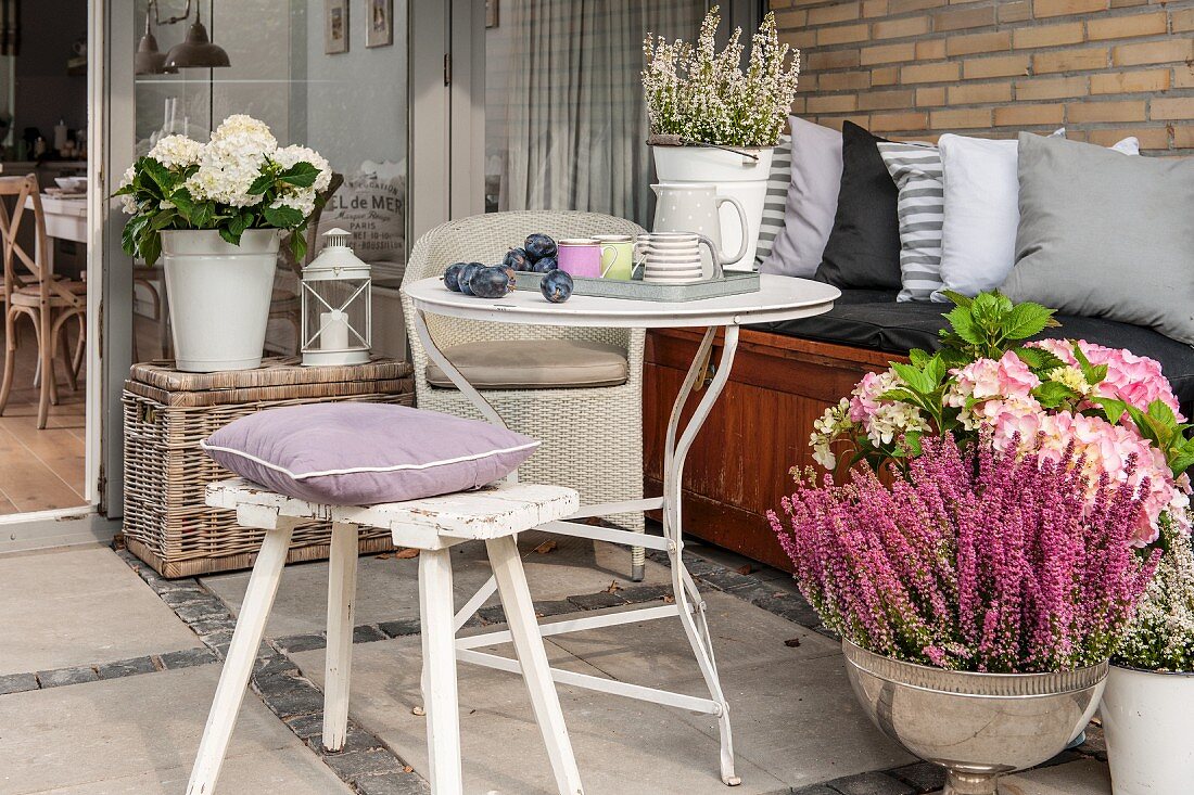 Kissen auf Hocker vor rundem Beistelltisch, neben bepflanzter Silberschale auf Terrasse