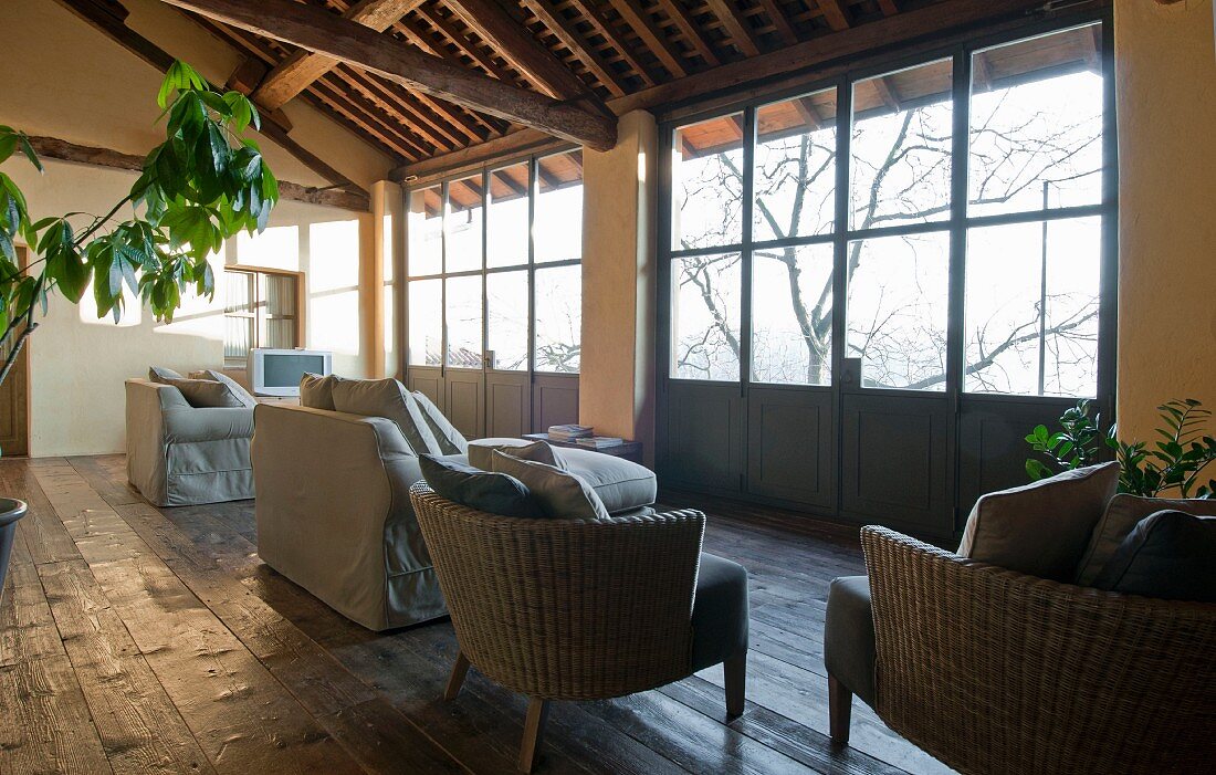 Wicker armchairs and sofas with pale grey loose covers on rustic wooden floor next to glazed terrace doors