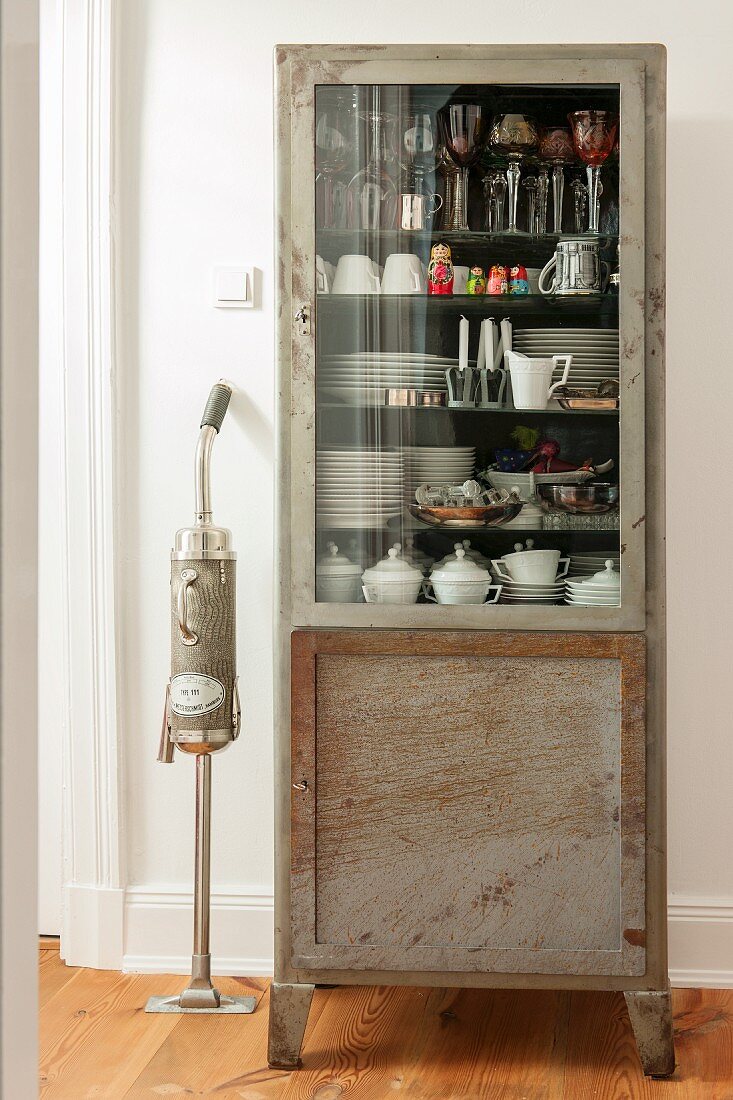 Retro vacuum cleaner next to crockery in vintage glass-fronted cabinet