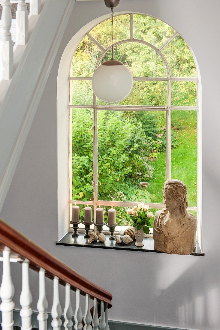 Bust of woman and candlesticks on sill of arched window with garden view in stairwell