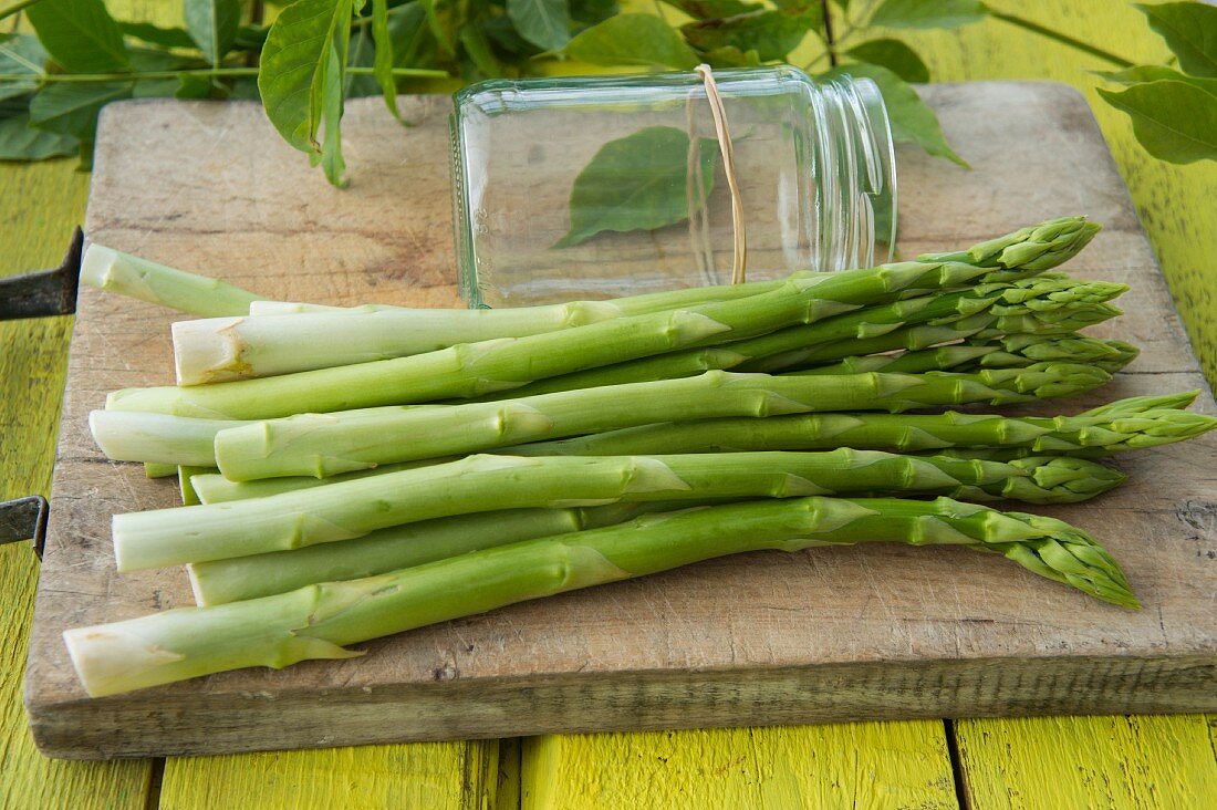 Grüner Spargel auf Holzschneidebrrett neben Schraubglas mit Gummi