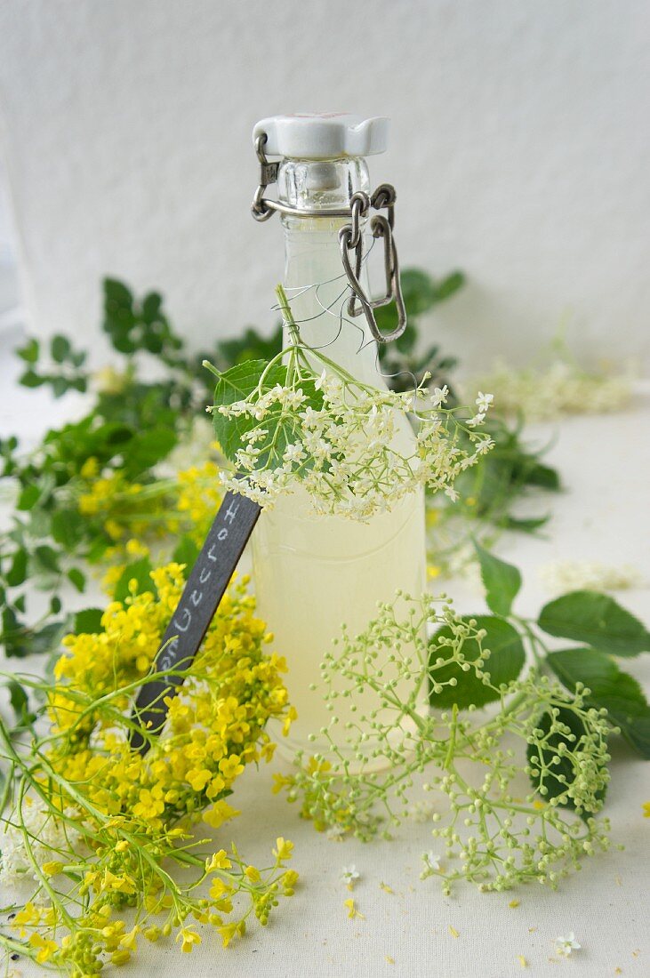 Elderflower syrup in a flip-top bottle, edlerflowers and a wreath of rape flowers