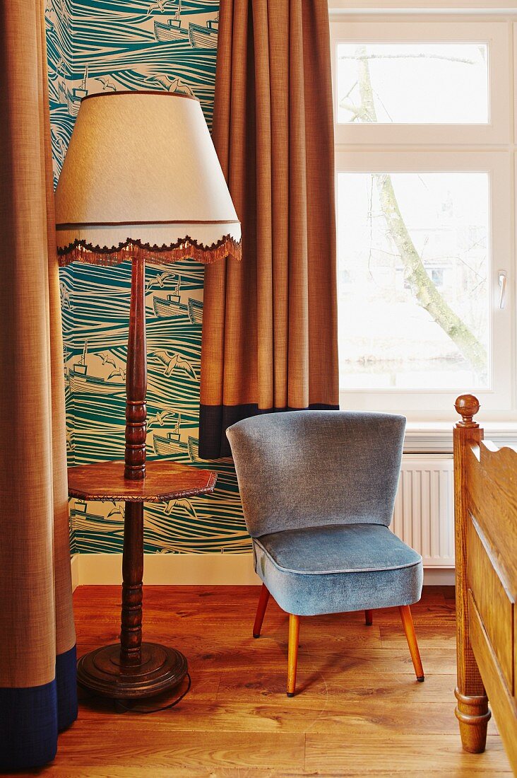 Sessel mit graublauem Samtbezug neben Stehleuchte mit Holzfuss und integrierter Ablage im Fiftiesstil in Zimmerecke