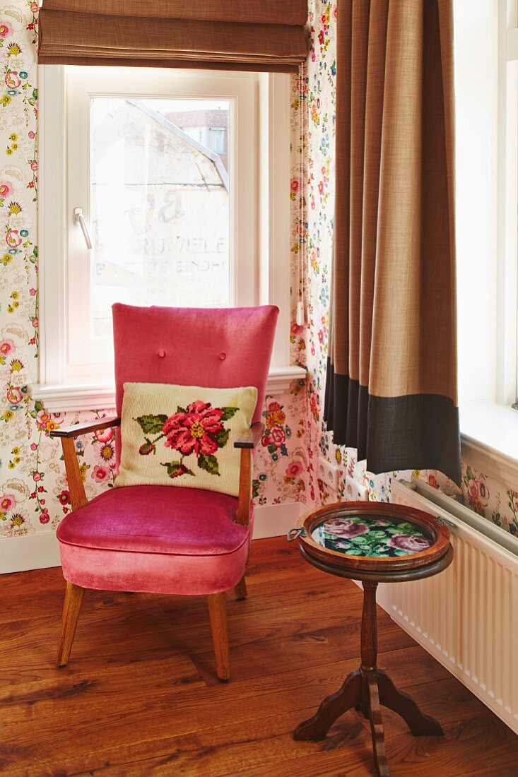 Kissen mit Blumenmotiv auf Sessel mit rosa Samtbezug neben zierlichem, antikem Beistelltisch aus Holz vor Fenster in Zimmerecke
