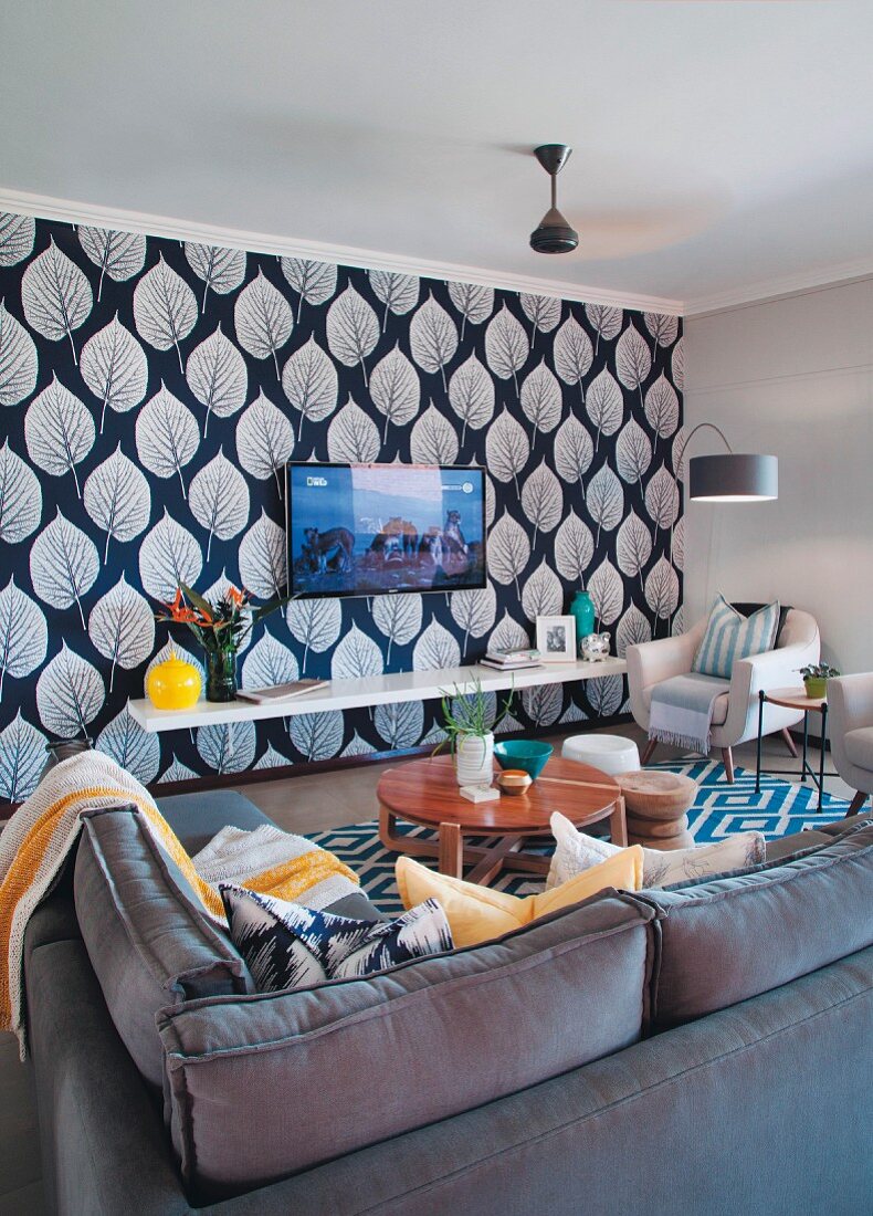 Living area with corner sofa and armchairs in front of TV mounted on wall with leaf-patterned wallpaper