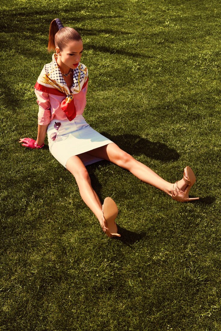 A young woman sitting on a lawn wearing a pink denim shirt and a white leather skirt
