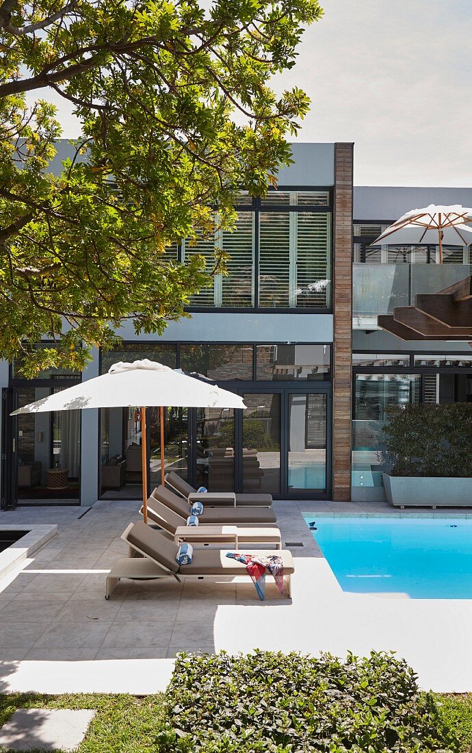 Sun loungers and parasol next to swimming pool in front of modern glass wall