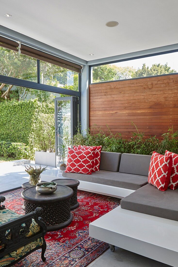 Lounge area with modern grey sofa, red and white scatter cushions and sliding glass wall leading to summery terrace