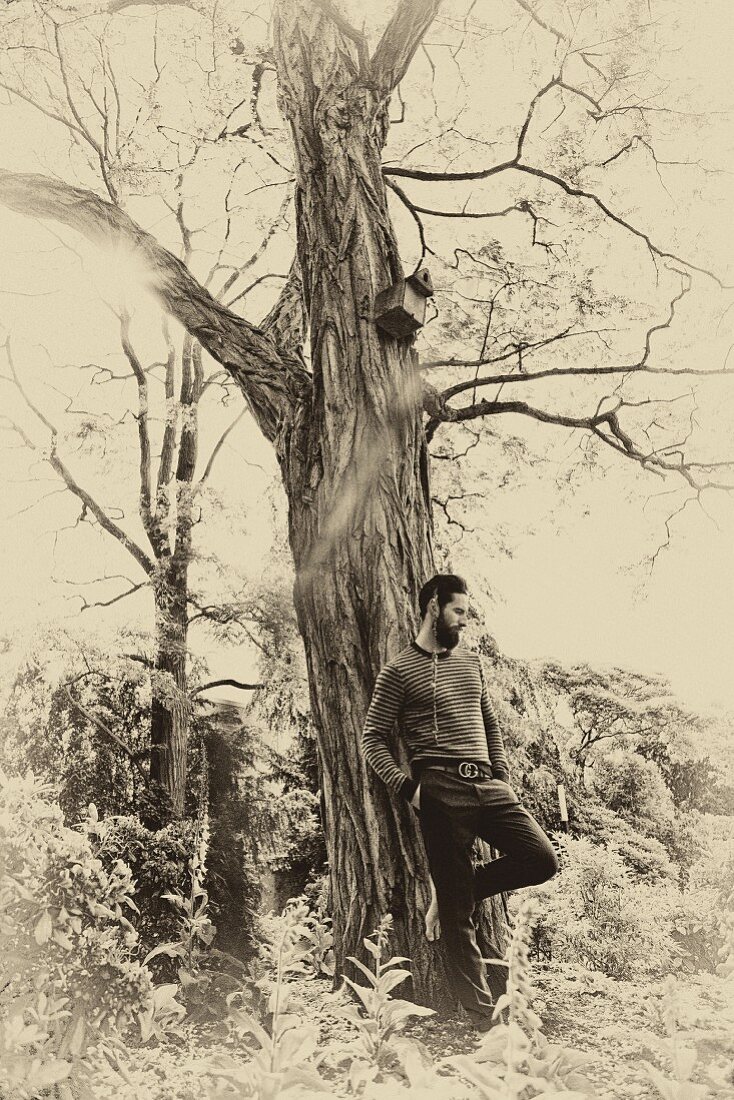 A man wearing woollen trousers and a striped knitted jumper leaning against an old tree