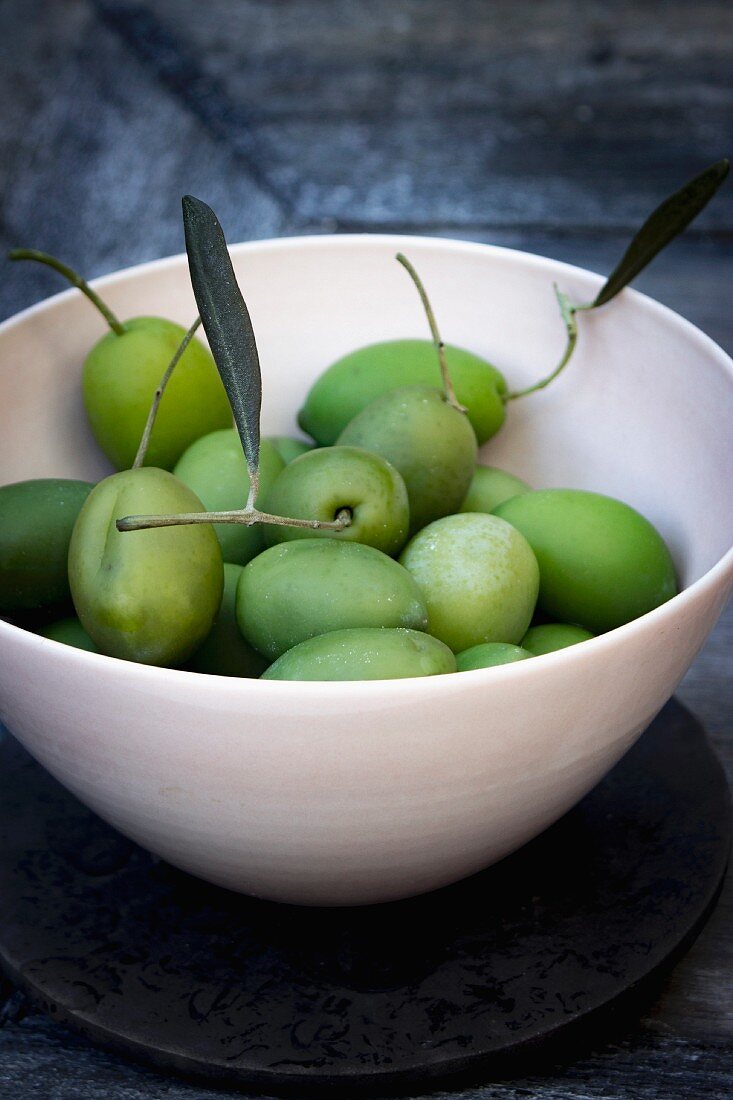 Grüne Oliven mit Stiel und Blättern in Schale auf grauem Holztablett