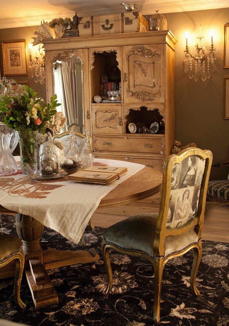 Antiker Stuhl mit vergoldetem Holzgestell und Vintage-Stoffdruck vor Tisch, im Hintergrund Holzschrank mit Spiegeltür