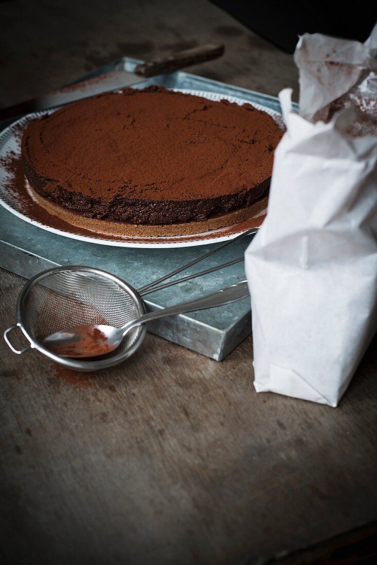 Chocolate tart on a metal tray with utensils for dusting cocoa powder on wooden table