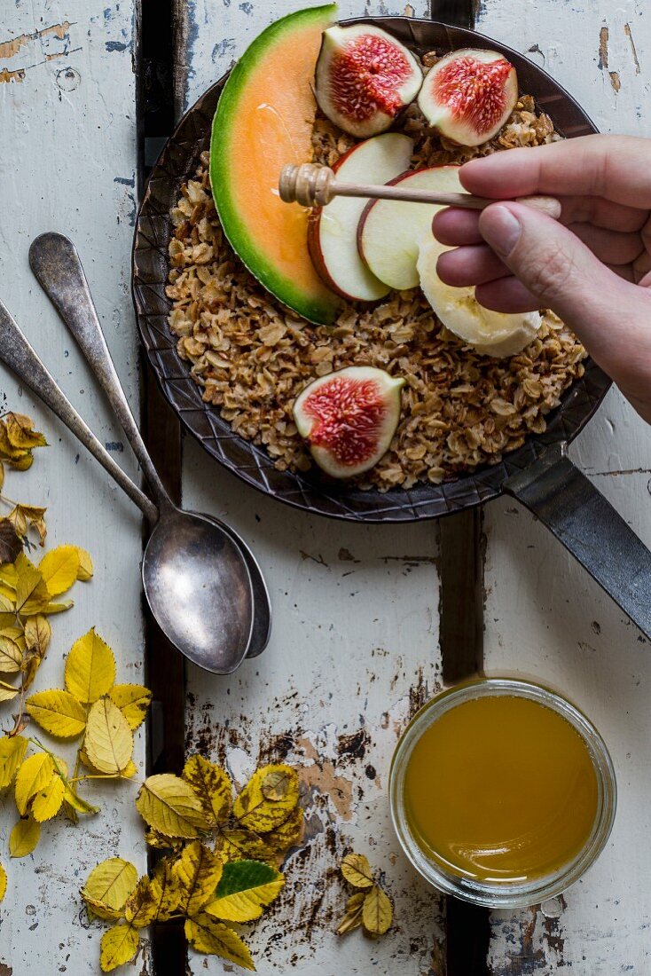 Honey being drizzled over roasted wholemeal oats in a cast-iron pan with fresh fruits
