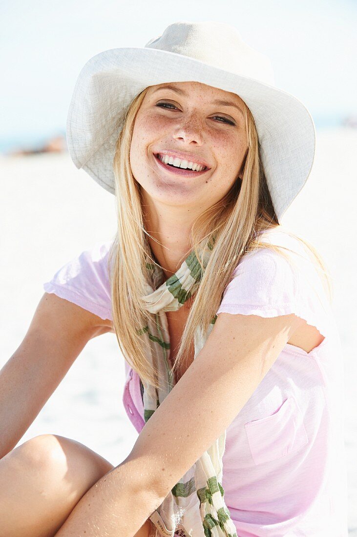 A young blonde woman on a beach wearing a summer hat, a scarf and a pink t-shirt