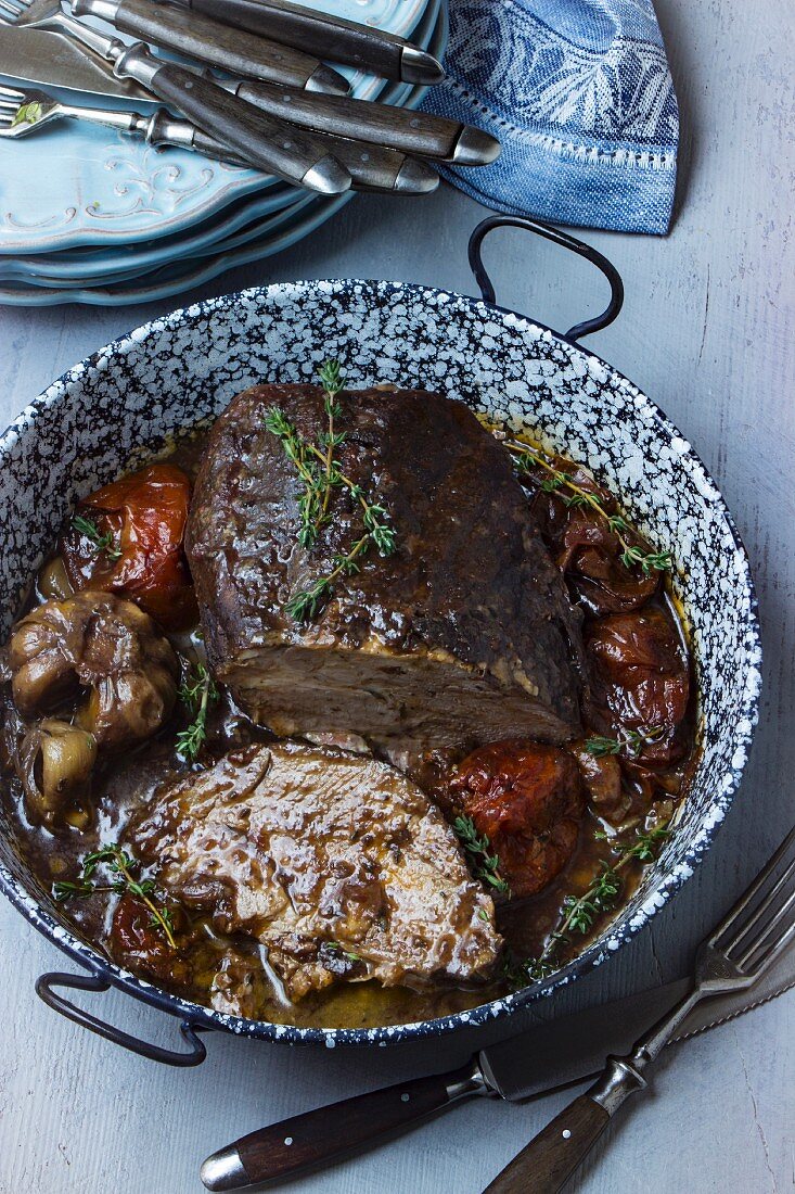 Italian roast pork with braised vegetables in a roasting tin with a stack of plates and cutlery