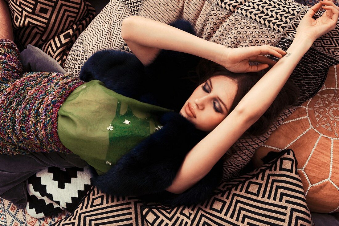A young woman wearing a blouse, a knitted skirt and a fur gilet lying on sofa cushions