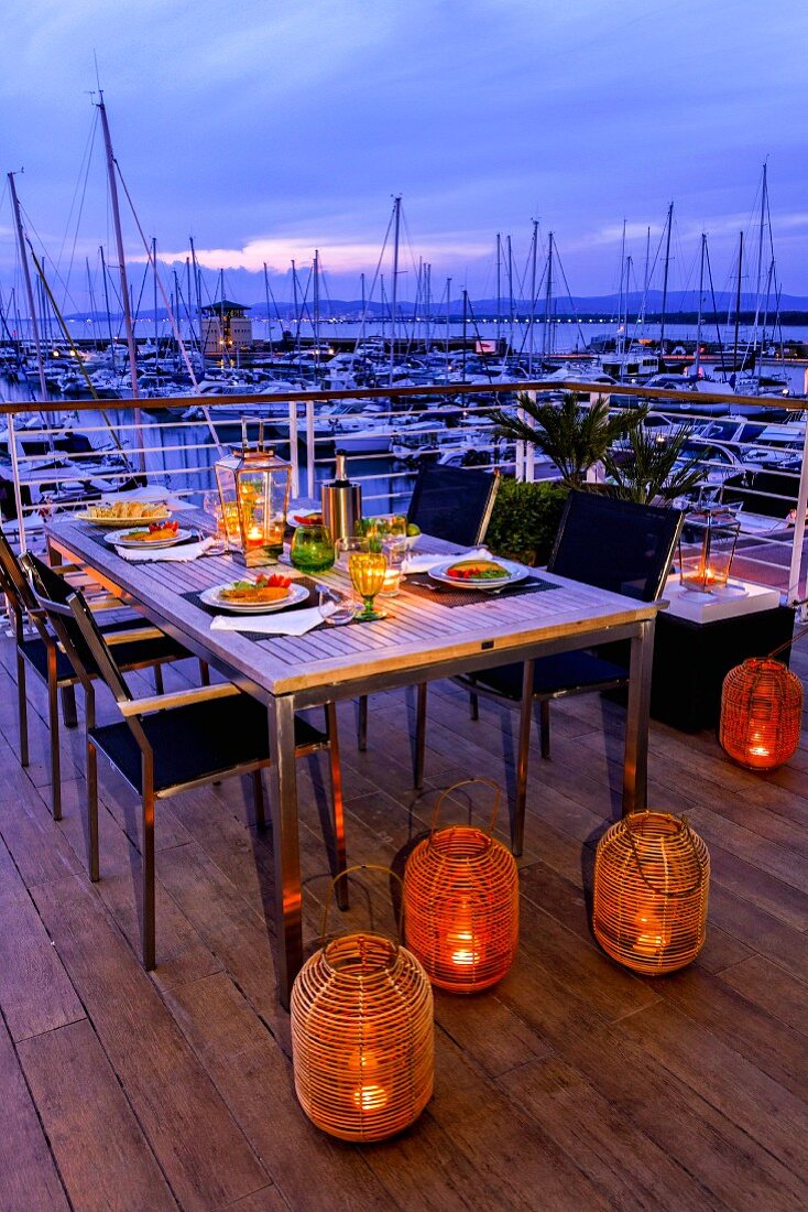 Bodenlaternen mit Kerzenlicht vor gedecktem Esstisch auf Terrasse, im Hintergrund Yachthafen in Abendstimmung